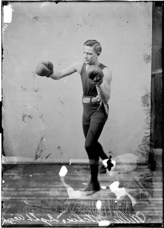 Pugilist, Willie Ritchie, facing image left, extending a right glove in a boxing stance in front of a light-colored backdrop - Chicago Daily News, Inc.
