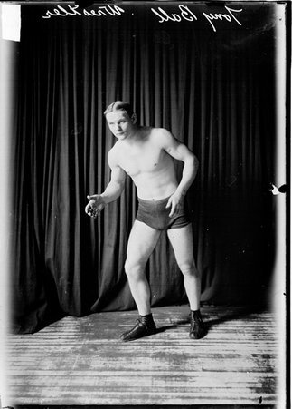 Wrestler, Tony Ball, standing with elbows bent and reaching forward slightly with right hand in front of a dark colored backdrop - Chicago Daily News, Inc.