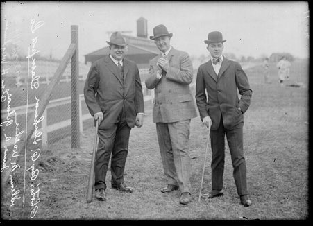Baseball player William M. Walker (clasping a baseball against his chest), James A. Gilmore, and Charles Weeghman standing in front of a long fence on a field - Chicago Daily News, Inc.