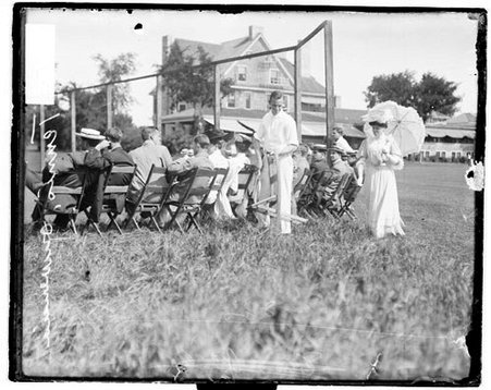 Unidentified man holding a wooden folding chair, walking with an unidentified woman on the side of a tennis court at Onwentsia Club - Chicago Daily News, Inc.
