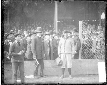 Baseball player Hugh Jennings, Detroit, carrying a bag, standing in front of a field seating area - Chicago Daily News, Inc.