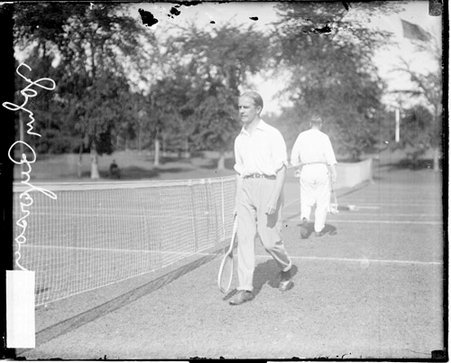 Tennis player John Ryerson holding a tennis racket, walking next to the net on a tennis court - Chicago Daily News, Inc.