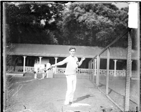 Tennis player Fred Carr holding a tennis racket out at his side next to a fence at the Aztec Tennis Club - Chicago Daily News, Inc.