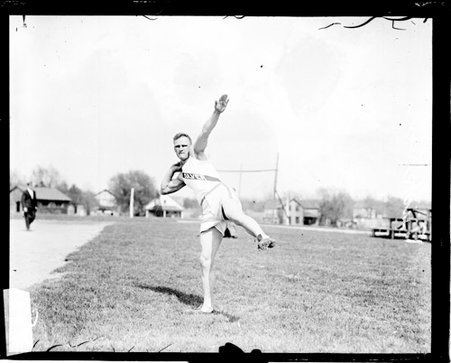 Athlete Dickson holding a shot put at shoulder-level and holding his left arm in the air, on an athletic field - Chicago Daily News, Inc.