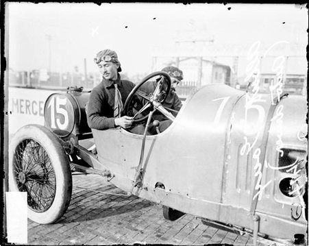 Automobile driver Goux sitting in the driver's seat of a parked automobile in front of a banner advertisement - Chicago Daily News, Inc.