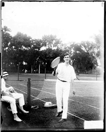 Tennis player Hayes holding a tennis racket in front of him, standing in front of the net on a tennis court - Chicago Daily News, Inc.