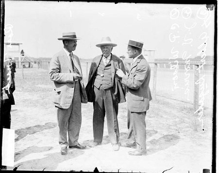 Racers C P Wilson, J V Morris, and Albert Luzader standing behind a low fence alongside a racetrack - Chicago Daily News, Inc.