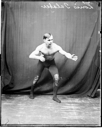 Wrestler, Louis Talaber leaning forward and extending his left arm in front of him in front of a dark backdrop in a room - Chicago Daily News, Inc.