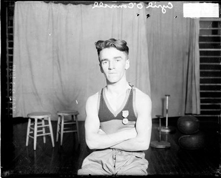 Wrestler, Cyril O'Connell sitting on stool with arms crossed in training room in front of light backdrop - Chicago Daily News, Inc.
