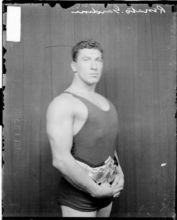 Wrestler, Renato Gardini standing in front of dark backdrop wearing trophy belt - Chicago Daily News, Inc.