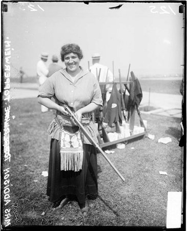 Shooter, Mrs. Topperwien standing in front of rack of rifles on lawn with men in background - Chicago Daily News, Inc.