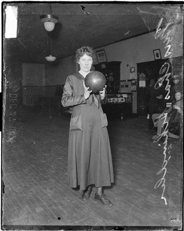Bowler, Mrs. Charles Schindler holding ball in front of her standing in room with men in background - Chicago Daily News, Inc.