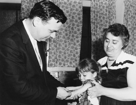 Mother looks on as young girl is being immunized by a shot administered by a doctor - Frank Las Studio