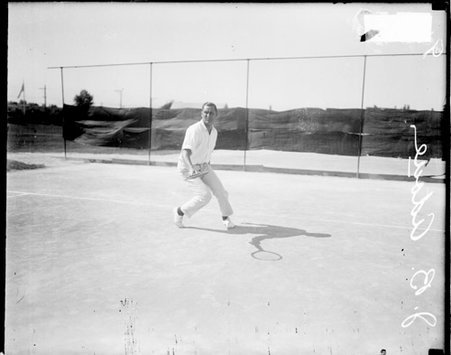 Tennis player, J. B. Adoue holding a tennis racket and leaning forward, standing on a tennis court - Chicago Daily News, Inc.