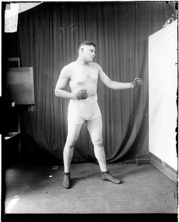 Boxer, Eddie Stock standing in profile in a boxing stance in front of a dark backdrop in a room - Chicago Daily News, Inc.