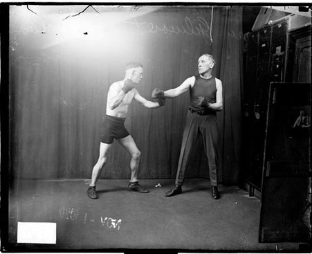 Pugilists, Gillmore and Douglas facing one another, standing in boxing stances in front of a dark backdrop in a room - Chicago Daily News, Inc.