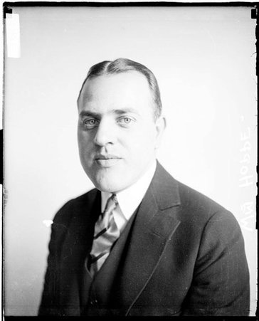 William Hoppe looking at the camera and wearing a suit, sitting in front of a light-colored backdrop in a room - Chicago Daily News, Inc.