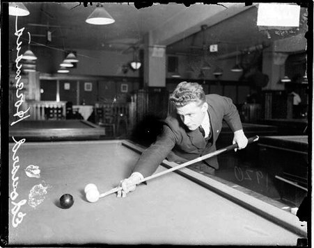 Billiards player, Horemans ready to hit a shot with a cue stick, standing next to a billiards table in a billiards hall in Chicago, Illinois. Billiards tables are visible in the background - Chicago Daily News, Inc.