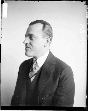 William Hoppe looking to the left of the image and wearing a suit, sitting in front of a light-colored backdrop in a room - Chicago Daily News, Inc.