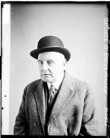 George G. Beltzhoover wearing a hat and street clothes, sitting in front of a light-colored backdrop in a room - Chicago Daily News, Inc.