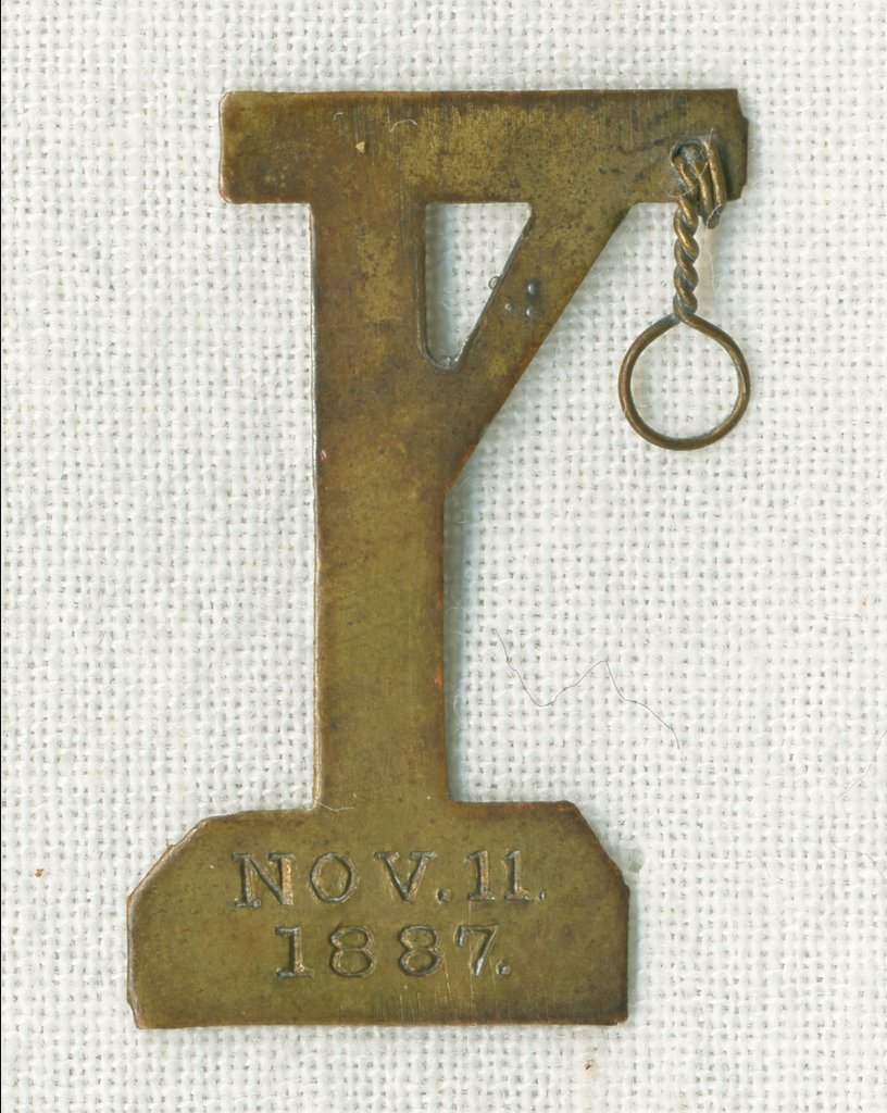 Brass lapel pin in the shape of a scaffold with a small wire noose attached to the top. Stamped into the metal at the bottom is Nov. 11, 1887. - Unknown